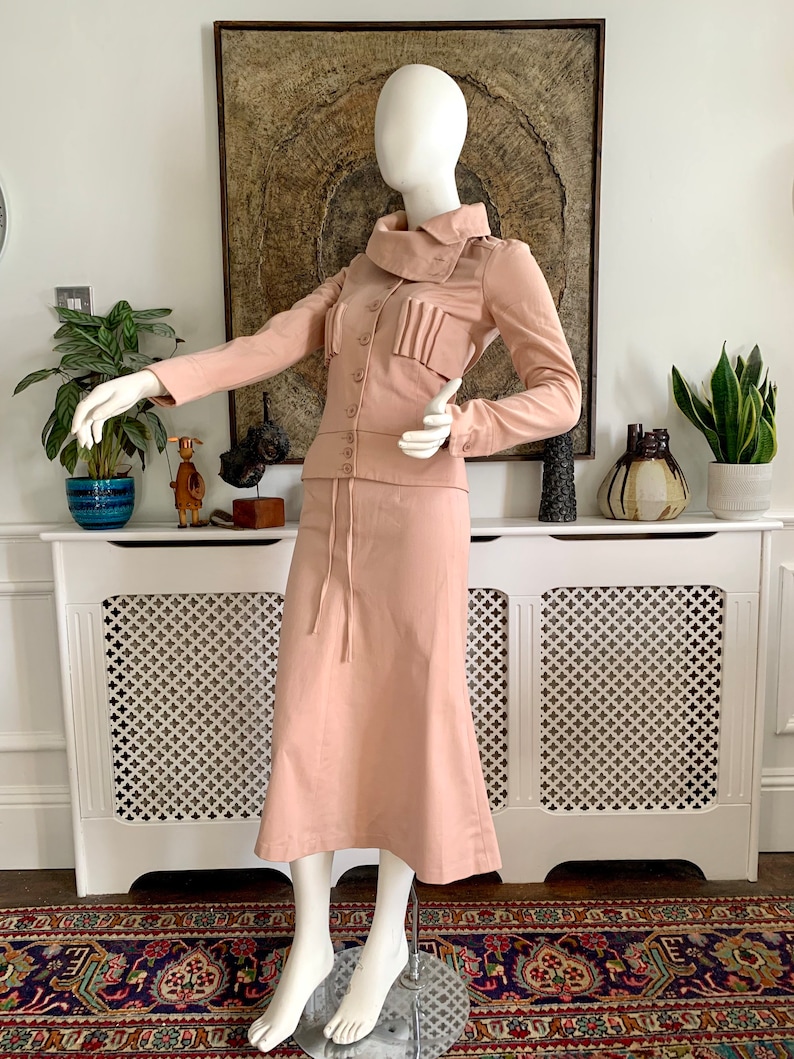 May include: A blush pink skirt suit with a long skirt and a fitted jacket featuring a high collar and a button closure. The jacket has a pleated detail on the front.