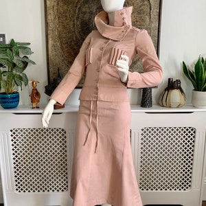 May include: A blush pink two-piece suit with a high collar jacket and a skirt with a drawstring waist.