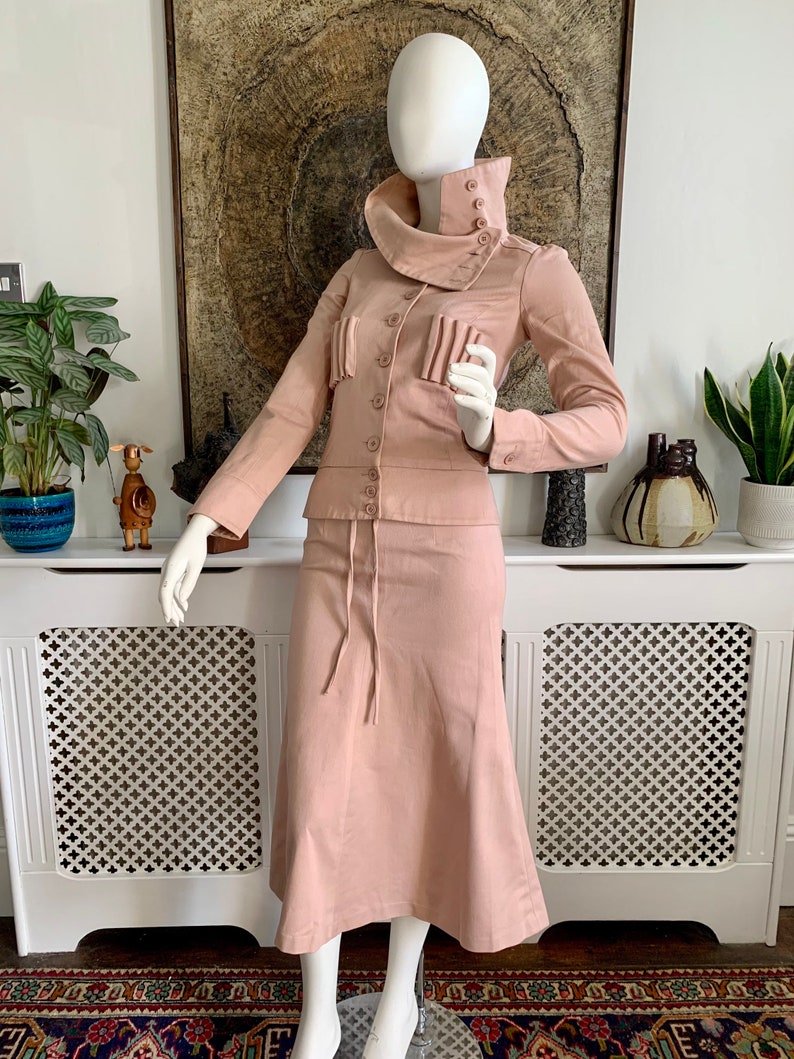 May include: A blush pink two-piece suit with a high collar jacket and a skirt with a drawstring waist.