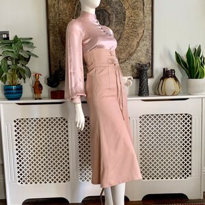 May include: A blush pink satin blouse with long sleeves and a high neck, paired with a blush pink high-waisted skirt with a lace-up detail at the front.