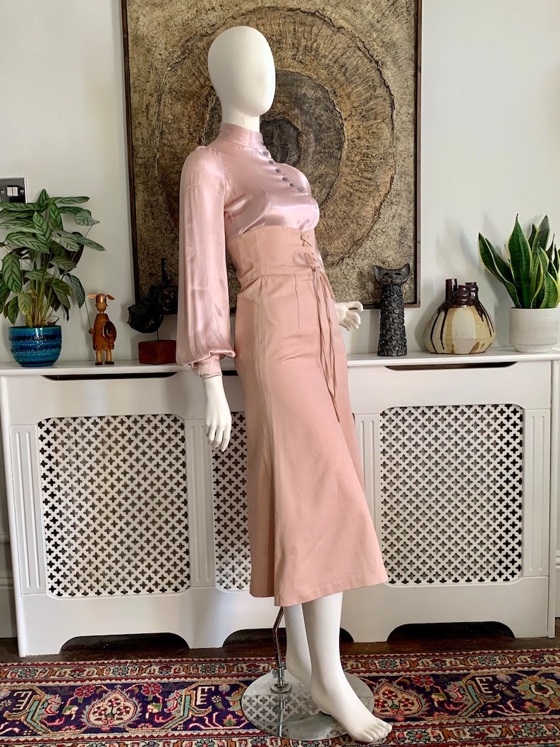 May include: A blush pink satin blouse with long sleeves and a high neck, paired with a blush pink high-waisted skirt with a lace-up detail at the front.
