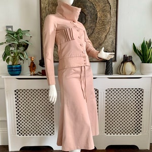 May include: A blush pink skirt suit with a high collar jacket and a skirt with a slight flare. The jacket has a button closure and a pleated detail on the front. The skirt has a drawstring waist.