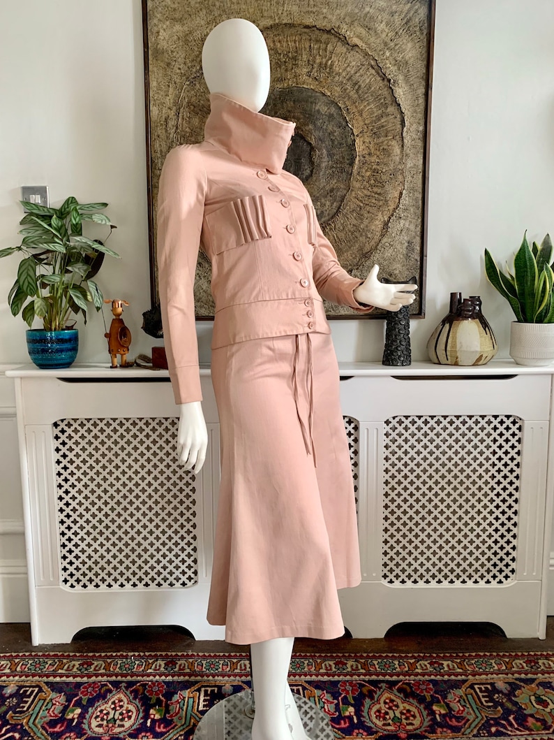 May include: A blush pink skirt suit with a high collar jacket and a skirt with a slight flare. The jacket has a button closure and a pleated detail on the front. The skirt has a drawstring waist.