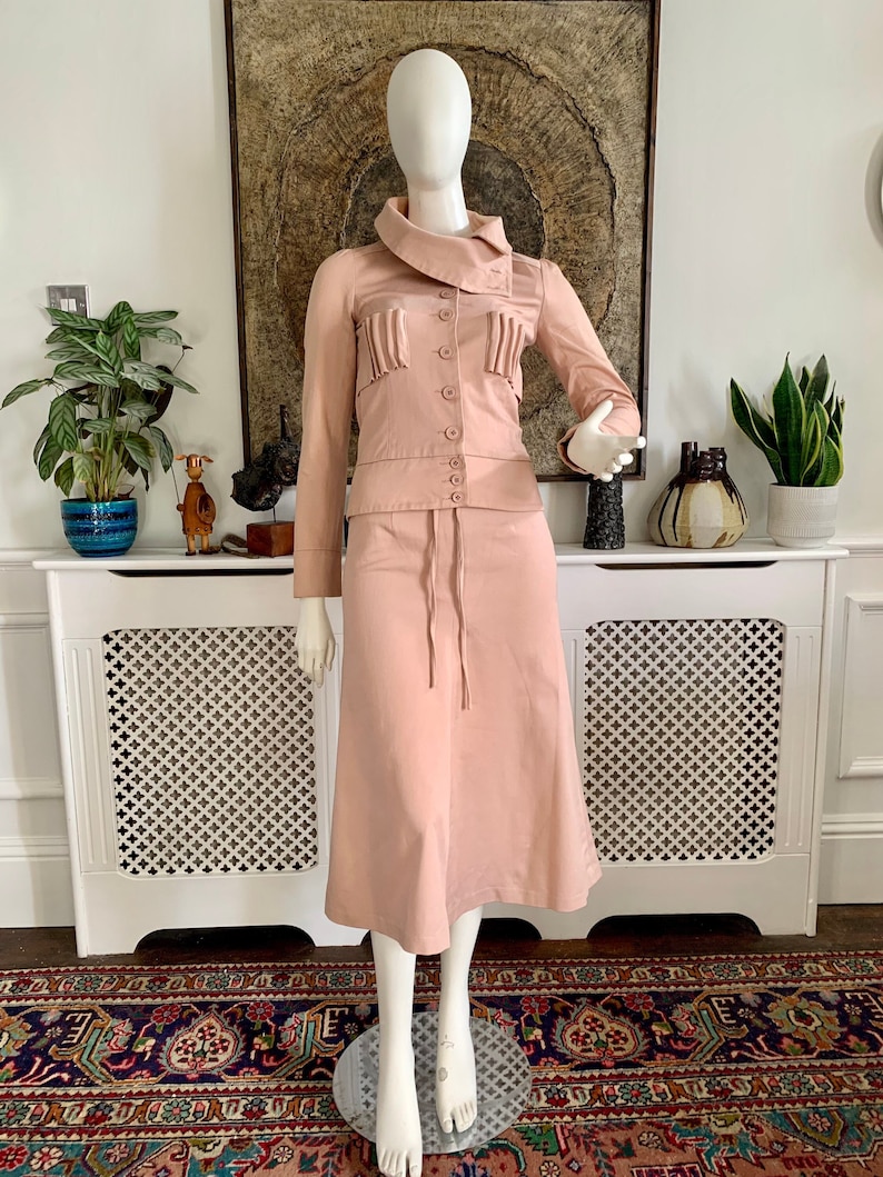 May include: A blush pink skirt suit with a long skirt and a fitted jacket. The jacket has a high collar and a button closure. The skirt has a drawstring waist and a flared hem.