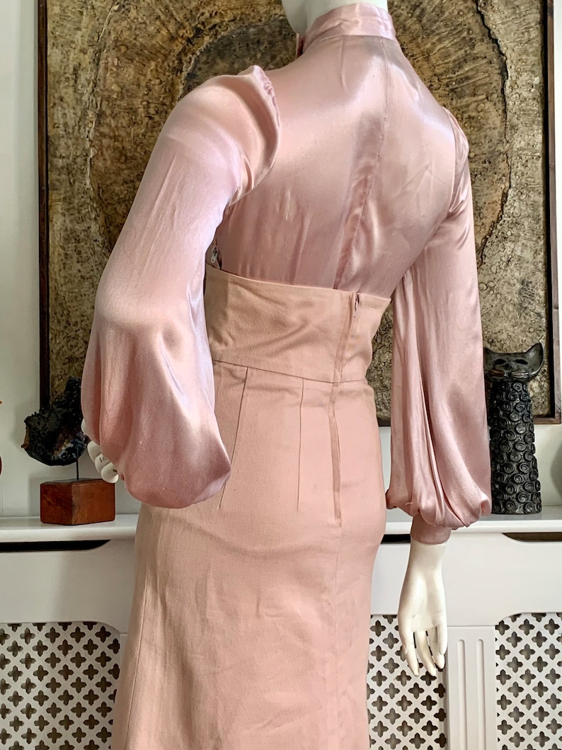 May include: A blush pink satin blouse with long sleeves and a high neck. The blouse is paired with a matching blush pink skirt that has a fitted waist and a straight skirt.
