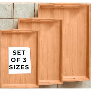 May include: A set of three bamboo serving trays in varying sizes. Each tray has a rectangular shape with a raised edge and a handle cutout. The text "SET OF 3 SIZES" is on a white label on the smallest tray.