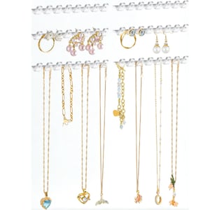 May include: A clear acrylic jewelry organizer displaying various gold necklaces, bracelets, rings, and earrings. The jewelry includes pieces with pearls, gemstones, and unique pendants. The organizer is designed to hang on a wall, showcasing the jewelry against a white background.