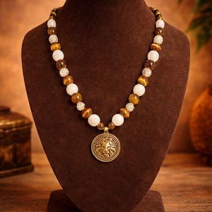 May include: A beaded necklace with a lion pendant displayed on a brown bust. The necklace features a mix of white, brown, and tiger-eye beads. The pendant is a gold-colored medallion with a detailed lion's head.