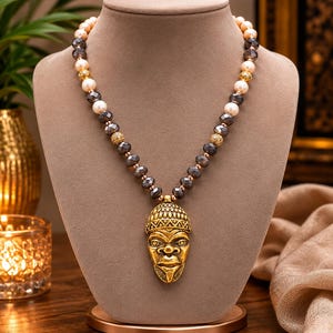 May include: A necklace featuring a gold-toned pendant shaped like a stylized human face with detailed facial features and a textured headdress. The strand consists of alternating cream-colored pearls, dark gray faceted beads, and small gold-toned spheres.