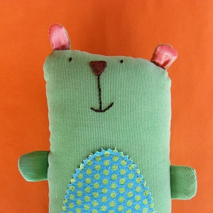 May include: Green corduroy stuffed animal shaped like a bear with a brown nose and black eyes. The bear has a blue and green polka dot pattern on its belly.