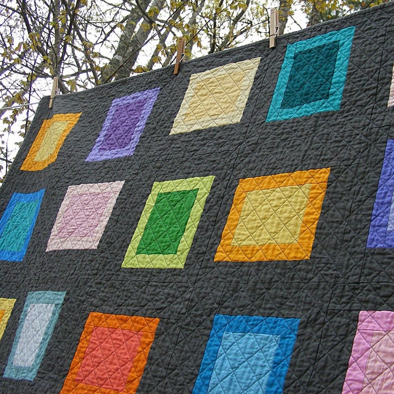 May include: A quilt with a gray background and colorful squares. The squares are in various colors, including pink, blue, green, yellow, and orange. The quilt is hanging on a clothesline.