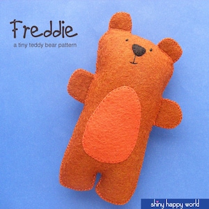 May include: A brown felt teddy bear with a red felt belly. The bear has black eyes and a stitched smile. The text "Freddie" and "a tiny teddy bear pattern" is above the bear. The text "shiny happy world" is below the bear.