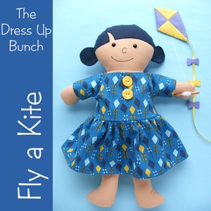 May include: A handmade doll wearing a blue and yellow dress with a diamond pattern. The doll is holding a purple and yellow kite with a string attached. The text "The Dress Up Bunch Fly a Kite" is on the left side of the image.