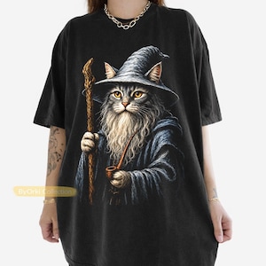 Peut inclure: Un t-shirt noir à manches courtes avec une impression graphique représentant un chat déguisé en magicien, portant un chapeau pointu et une robe sombre, tenant un bâton en bois et une pipe. Le texte ByOrki Collection est imprimé en jaune.