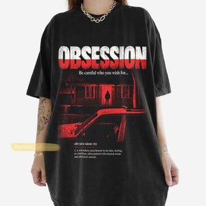 May include: A black short-sleeve t-shirt featuring a red and white graphic print. The design includes the text "OBSESSION", "Be careful who you wish for...", and a dictionary definition of obsession above a dark house and car illustration.