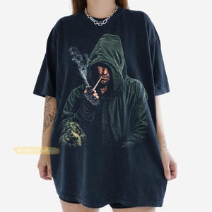 May include: A black short-sleeve t-shirt featuring a graphic print of a hooded figure in a dark green cloak smoking a pipe with visible smoke. The figure wears dark gloves and has a textured appearance. The text ByOrki Collection is visible.