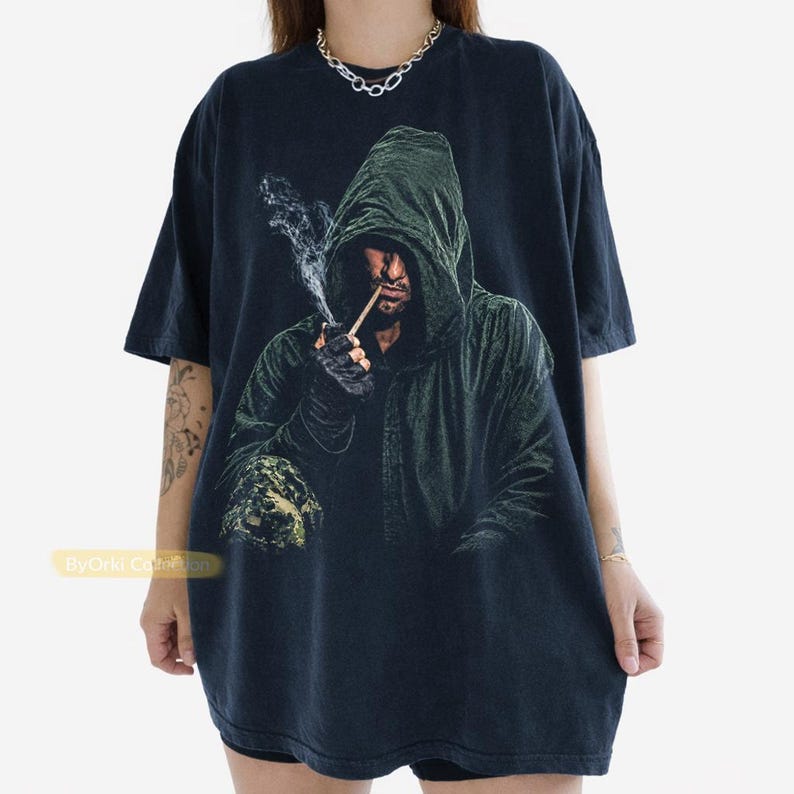 May include: A black short-sleeve t-shirt featuring a graphic print of a hooded figure in a dark green cloak smoking a pipe with visible smoke. The figure wears dark gloves and has a textured appearance. The text ByOrki Collection is visible.