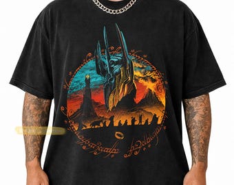 May include: A black short-sleeve t-shirt featuring a graphic print of a dark armored helmet, a volcanic landscape, and a silhouette of a group of figures. The design is encircled by a ring with glowing orange text and a small ring icon at the bottom.