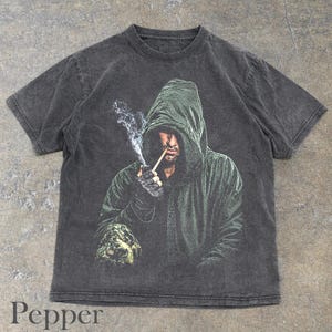 May include: A charcoal gray short-sleeve t-shirt featuring a graphic print of a hooded figure in a dark green cloak. The figure is holding a lit cigarette with visible smoke rising. The shirt has a faded, washed-out texture throughout the fabric.