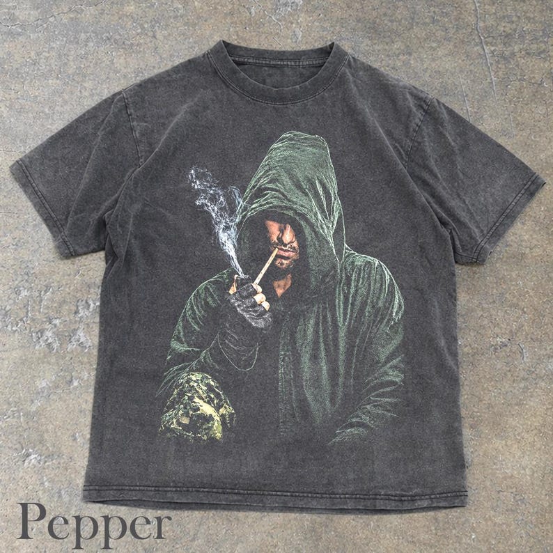 May include: A charcoal gray short-sleeve t-shirt featuring a graphic print of a hooded figure in a dark green cloak. The figure is holding a lit cigarette with visible smoke rising. The shirt has a faded, washed-out texture throughout the fabric.