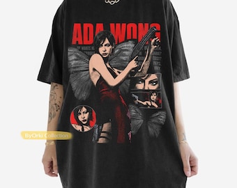 May include: A black short-sleeve t-shirt featuring a graphic print of Ada Wong with butterfly wings, holding a firearm. The design includes the text ADA WONG in bold red letters, smaller inset images of the character, and additional text elements.