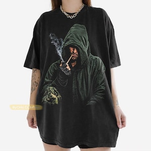 Peut inclure: Un t-shirt noir à manches courtes avec une impression graphique représentant une personne portant une cape à capuche vert foncé, fumant une pipe avec de la fumée visible. La personne porte des gants sombres et a une expression sérieuse.