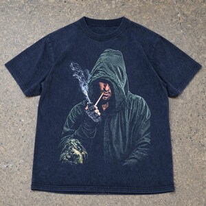May include: A dark charcoal gray short-sleeve t-shirt featuring a graphic print of a hooded figure in a dark green cloak. The figure is shown smoking a thin pipe with visible wisps of white smoke rising upward against the dark fabric background.