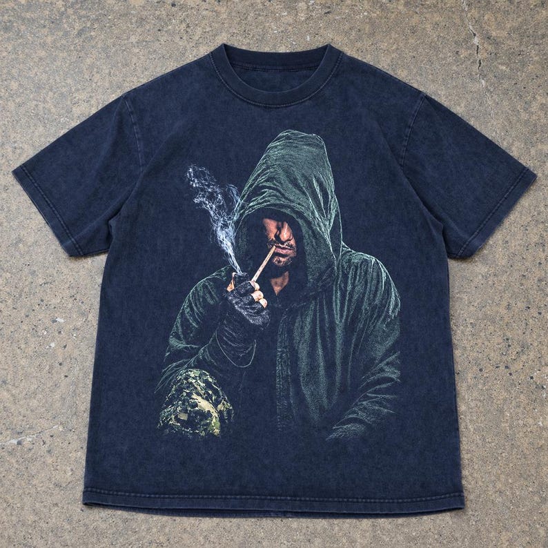 May include: A dark charcoal gray short-sleeve t-shirt featuring a graphic print of a hooded figure in a dark green cloak. The figure is shown smoking a thin pipe with visible wisps of white smoke rising upward against the dark fabric background.