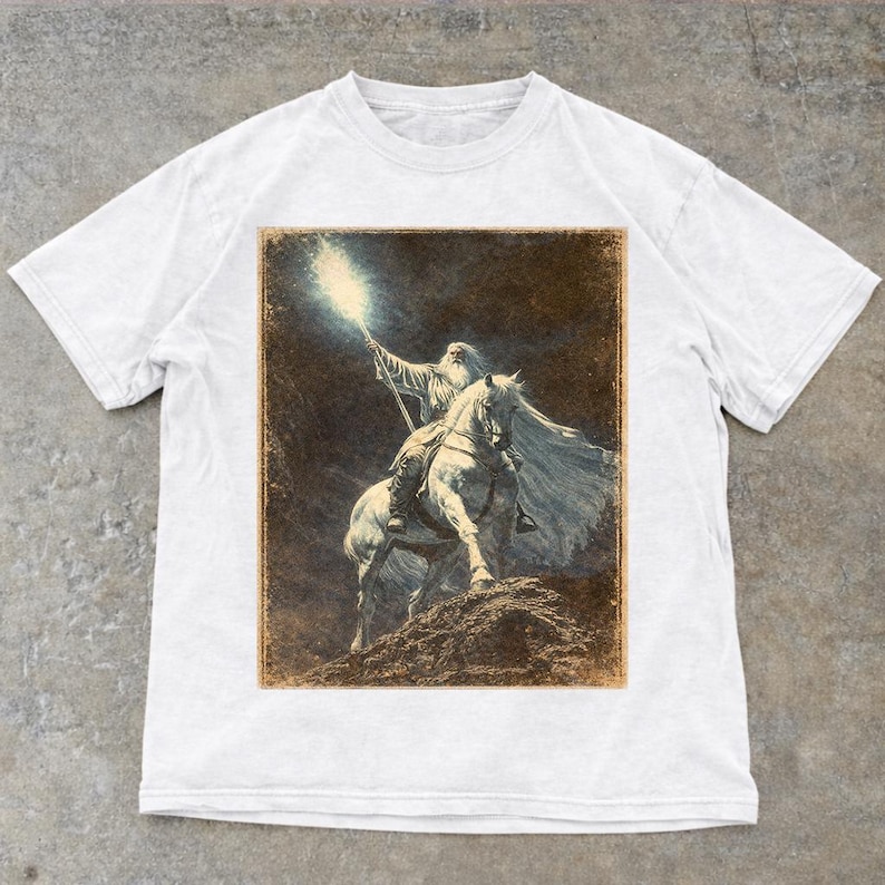 White Vintage Gandalf on horseback with glowing staff in a vintage fantasy illustration, lord of the rings inspired wizard design, lotr fan art style graphic tee, magical wizard scene with detailed horse and mystical light, fantasy lover gift, wizard gift idea, lord of the ring shirt, gandalf shirt, fantasy aesthetic clothing, unisex streetwear tee, high quality print, perfect gift for lotr fans and magic lovers