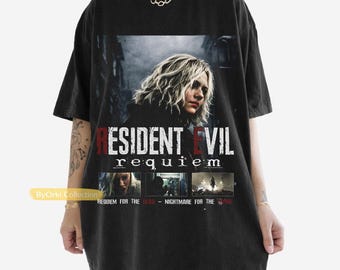 May include: A black short-sleeve t-shirt featuring a graphic print of a person with blonde hair. The text reads RESIDENT EVIL requiem, with smaller images below and the text REQUIEM FOR THE DEAD - NIGHTMARE FOR THE LIVING.