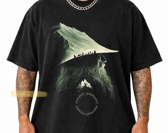 May include: A black short-sleeve t-shirt featuring a graphic print of a bearded figure with a mountain-shaped hat. A line of small silhouettes walks along the hat's brim, while a circular ring with elvish script is positioned below the beard.