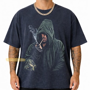 May include: A charcoal gray short-sleeve t-shirt featuring a graphic print of a hooded figure in a dark green cloak smoking a cigarette. The figure wears dark gloves and is partially obscured by shadow. The shirt has a faded, vintage-style wash.