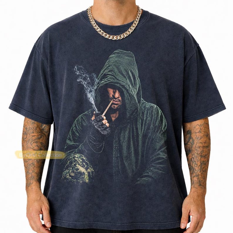 May include: A charcoal gray short-sleeve t-shirt featuring a graphic print of a hooded figure in a dark green cloak smoking a cigarette. The figure wears dark gloves and is partially obscured by shadow. The shirt has a faded, vintage-style wash.