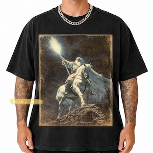 Puede incluir: Camiseta negra con una ilustración vintage de un mago a caballo, sosteniendo un bastón que emite luz brillante. La obra de arte tiene un tono sepia, con una representación detallada del mago y el caballo. La camiseta tiene un cuello redondo clásico.