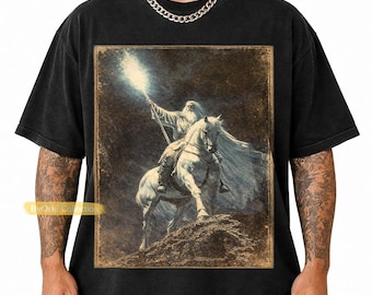 May include: Black t-shirt featuring a vintage-style illustration of a wizard on horseback, holding a staff emitting bright light. The artwork has a sepia tone, with a detailed depiction of the wizard and horse. The shirt has a classic crew neck.