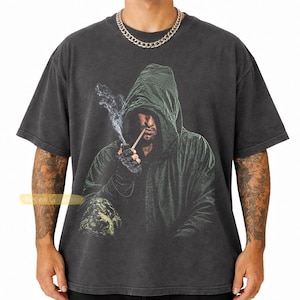 May include: A charcoal gray short-sleeve t-shirt featuring a graphic print of a hooded figure in a dark green cloak smoking a pipe. The figure wears dark gloves and is surrounded by wisps of smoke. The shirt has a crew neckline and a relaxed fit.