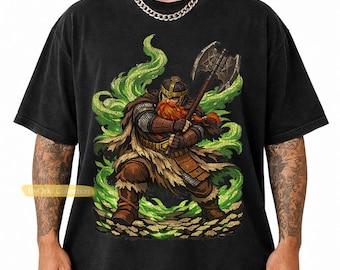 May include: A black short-sleeve t-shirt featuring a graphic print of a bearded warrior in armor holding a large battle axe. The warrior is surrounded by swirling green energy effects and stands on a rocky ground. The text ByOrki Collection is visible.