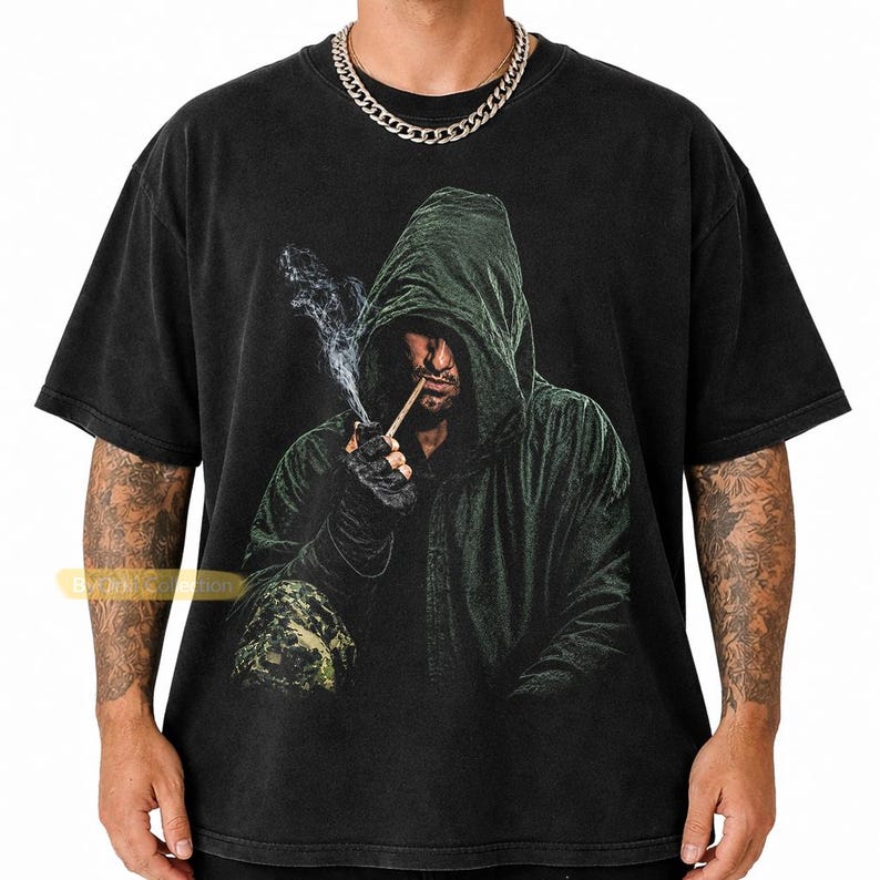 May include: A black short-sleeved t-shirt featuring a graphic print of a hooded figure in a dark green cloak smoking a pipe. The figure wears dark gloves and has a camouflage pattern on the lower portion of the garment. The shirt has a crew neck.