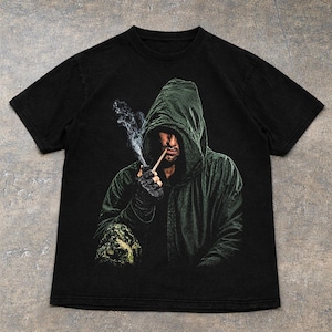 May include: A black short-sleeve t-shirt featuring a graphic print of a person wearing a dark green hooded cloak. The person is holding a smoking pipe to their mouth, with visible wisps of smoke rising upward against the dark background.