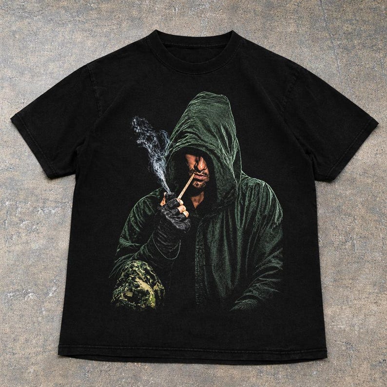 May include: A black short-sleeve t-shirt featuring a graphic print of a person wearing a dark green hooded cloak. The person is holding a smoking pipe to their mouth, with visible wisps of smoke rising upward against the dark background.