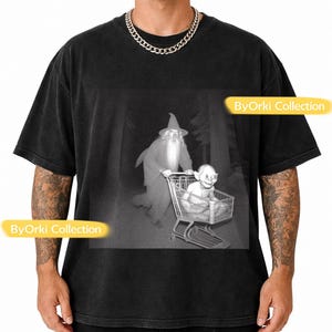 Puede incluir: Camiseta negra con un gráfico en blanco y negro de Gandalf empujando un carrito de la compra con Gollum. La escena está ambientada en un bosque oscuro. El texto "ByOrki Collection" está en la camiseta.