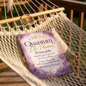 May include: A purple and gold "Quantum Life Planner Printable" journal rests on a white hammock. The journal includes text for manifestation, vision boards, and affirmations. A dreamcatcher hangs above.
