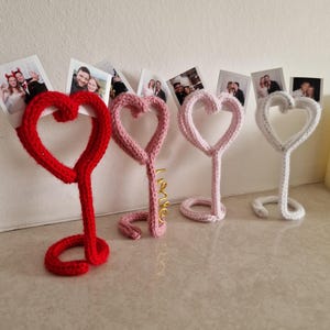 May include: Four heart-shaped photo holders in red, pink, light pink, and white. The holders are made of a knitted material and have a looped base. The pink holder has the word "love" in gold wire. Photos are displayed above the holders.
