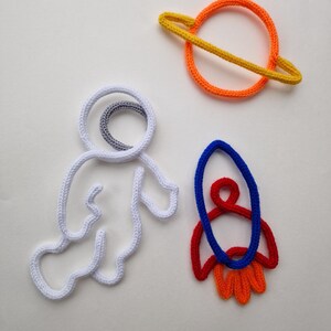 May include: Handmade crochet space-themed decorations. Includes a white astronaut, a planet with orange and yellow rings, and a red, blue, and orange rocket. The items are made with thick yarn and are set against a white background.