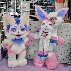 May include: Two plush animal figures. The left figure is white with purple and pink accents, large ears, and red eyes. The right figure is white and pink with long ears, blue eyes, a large white bow, and a pink glittery bow tie with ribbon streamers.