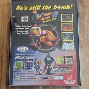 May include: Framed poster for the Nintendo 64 game, Bomberman 64: The Second Attack! The poster features the game's logo, character illustrations, and gameplay screenshots. The text "He's still the bomb!" is at the top.