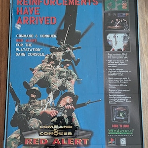 May include: A framed video game box for Command & Conquer: Red Alert for the PlayStation. The cover features soldiers, a helicopter, and the text "REINFORCEMENTS HAVE ARRIVED." The game's title is prominently displayed.