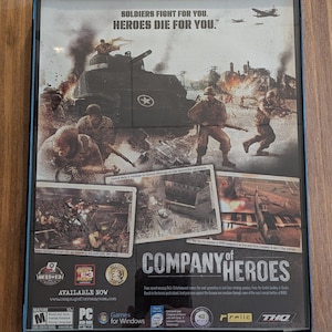 以下が含まれることがあります： 「Company of Heroes」のフレーム入りビデオゲームケース。兵士、戦車、爆発のある戦争シーンが描かれています。ケースには「SOLDIERS FIGHT FOR YOU. HEROES DIE FOR YOU.」と「AVAILABLE NOW」の文字が含まれています。