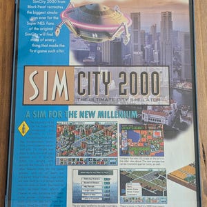 May include: SimCity 2000 video game box for Super NES. The box art features a futuristic cityscape with a flying saucer and the game's title. Includes screenshots and text describing gameplay features.