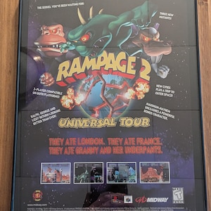 Pode incluir: Pôster emoldurado do videogame Rampage 2 Universal Tour. O pôster apresenta três monstros, Ralph, George e Lizzie, com o texto "They ate London. They ate France. They ate Granny and her underpants."